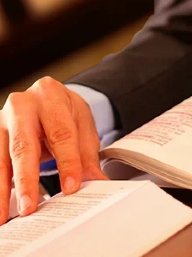 Story Imagem de um homem com a mão apoiada sobre livros e documentos em uma mesa, simbolizando estudo e dedicação aos estudos.