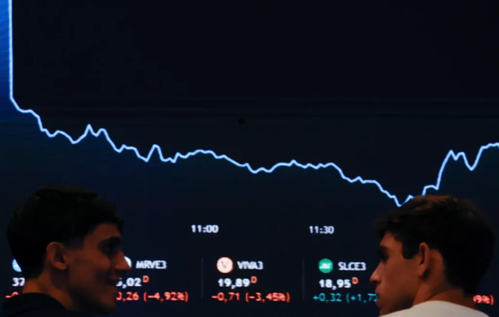 Dois jovens observando gráficos de ações em uma tela, representando o movimento do mercado financeiro e suas flutuações ao longo do tempo.