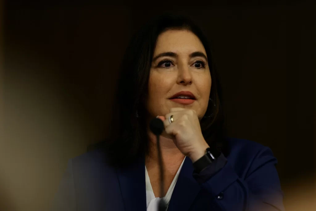 Imagem de uma mulher em evento público, com expressão confiante e gestos enfáticos. Ela está usando um blazer azul e segurando um microfone, aparentemente durante um discurso.