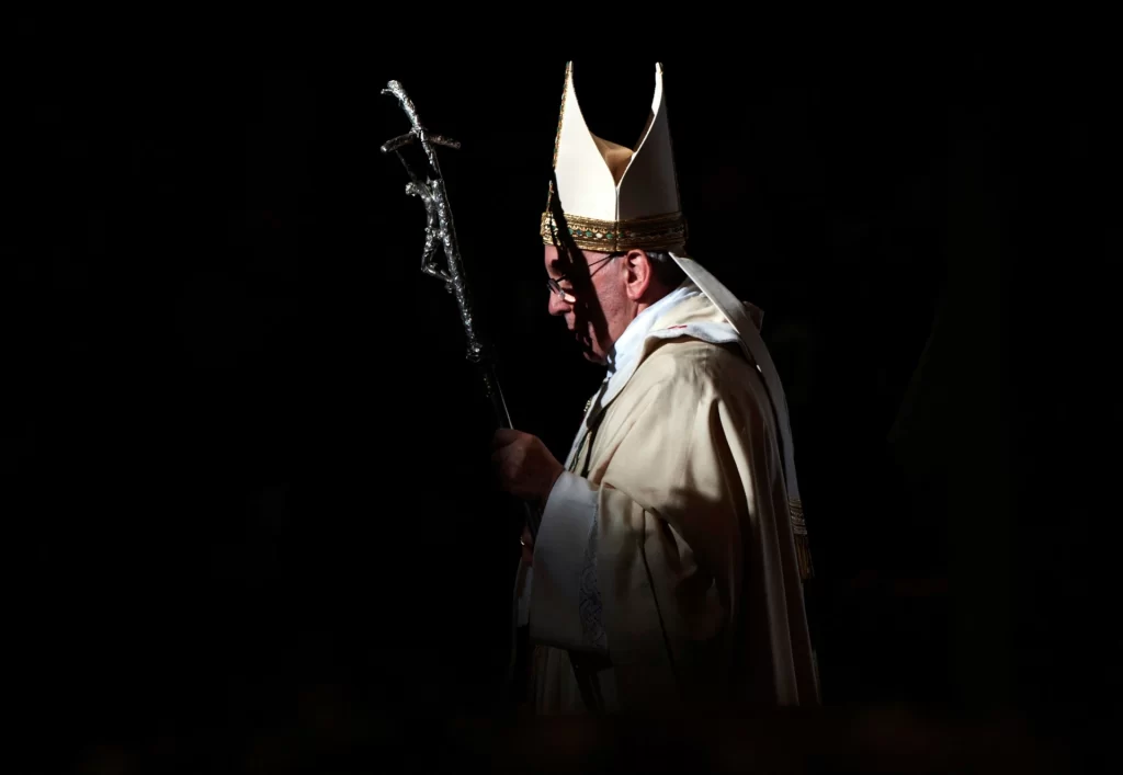 Imagem do Papa Francisco durante uma cerimônia religiosa, vestido com trajes litúrgicos, segurando um cajado, em um ambiente soturno e rezando.
