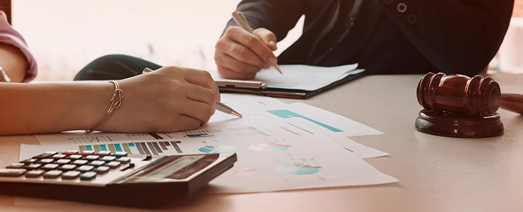 Mãos de duas pessoas analisando documentos financeiros e gráficos sobre uma mesa, com uma calculadora e um martelo de juiz ao fundo.