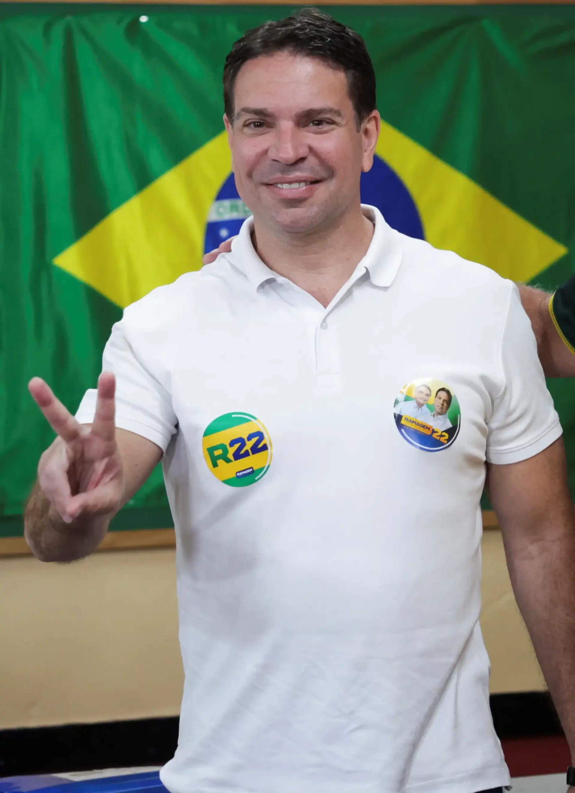 Homem sorridente com camiseta branca e bottons de campanha eleitoral, fazendo sinal de paz em frente a bandeira do Brasil.