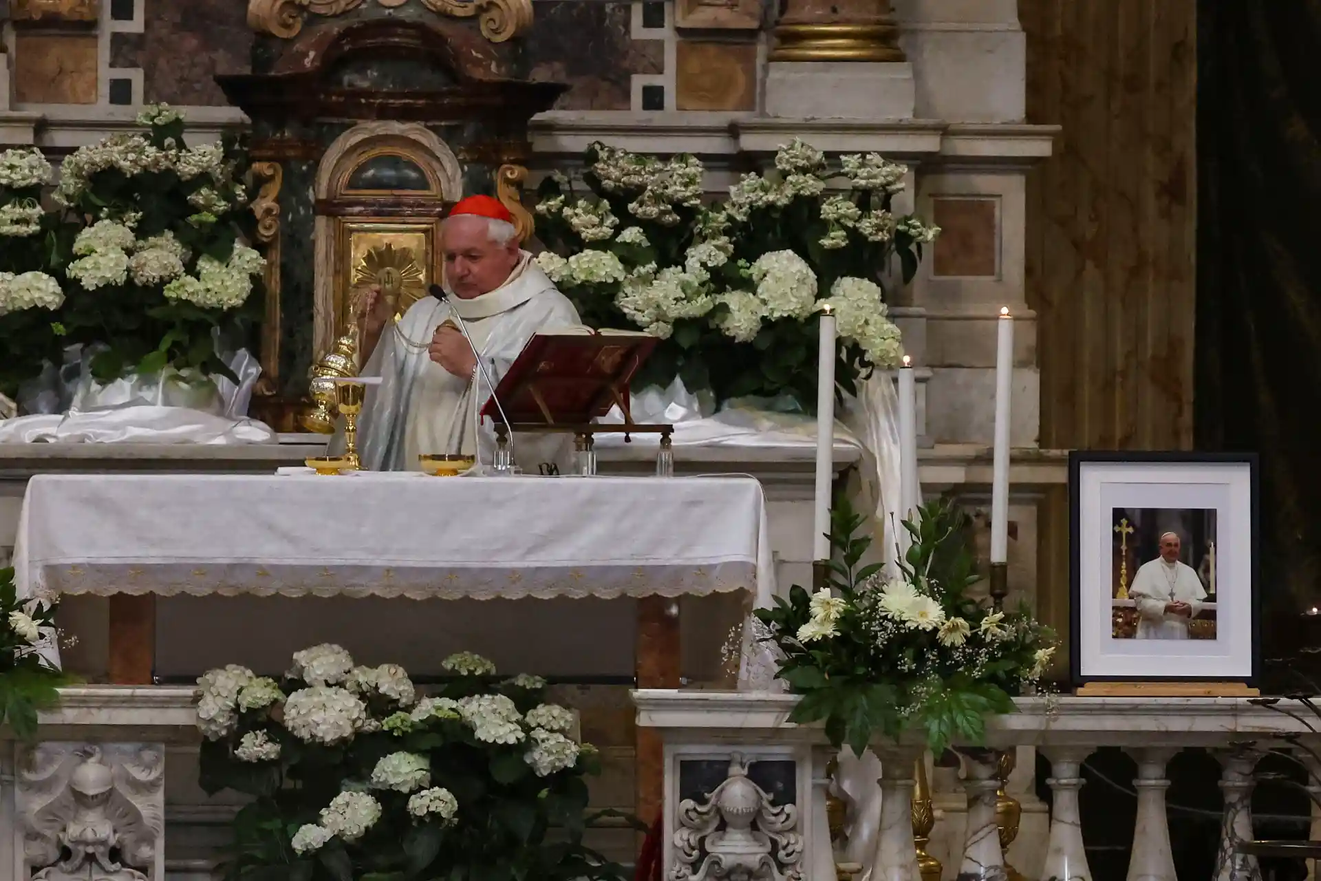 Um cardeal oferecendo uma missa em um altar decorado com flores brancas e velas em um ambiente religioso.