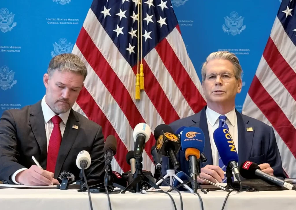 Imagem de uma conferência de imprensa, com dois homens em traje formal, uma bandeira americana ao fundo, e microfones na mesa, representando diplomacia.