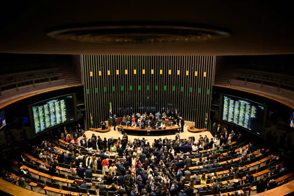 Imagem do Congresso Nacional do Brasil durante uma sessão com muitos deputados e funcionamento dos painéis eletrônicos, simbolizando a democracia e a atividade legislativa.