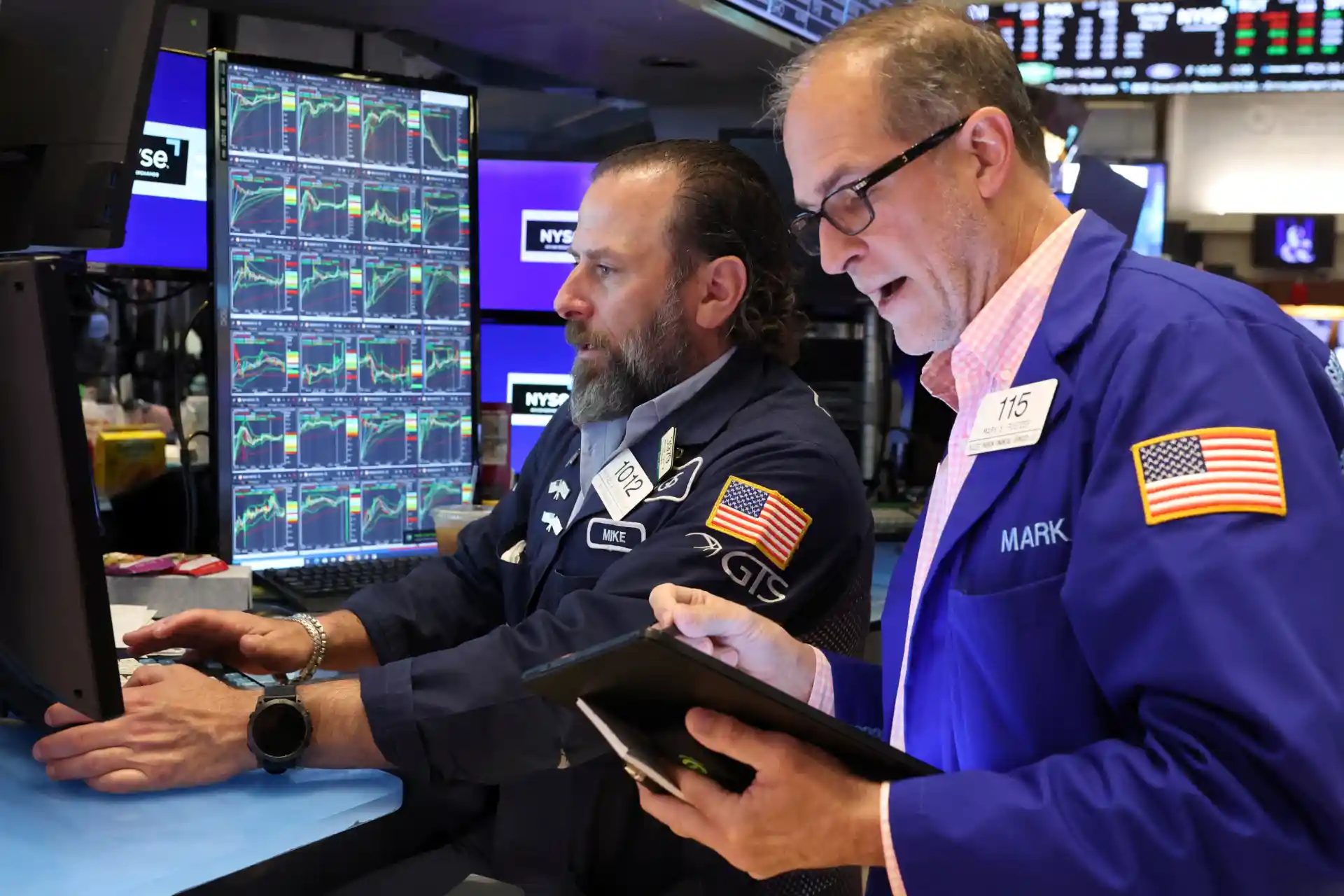 Dois corretores analisando gráficos no pregão da bolsa de valores, um deles utilizando tablet e o outro um computador. Ambiente financeiro intenso e tecnológico.
