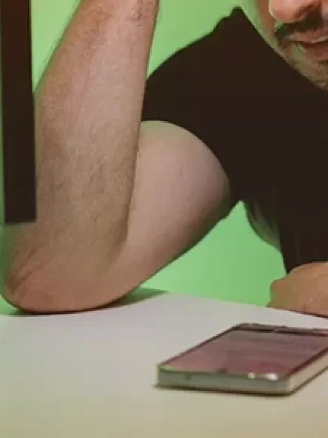 Story Homem pensativo olhando para o celular em uma mesa, com um computador ao fundo e iluminação verde. Situação de stress ou concentração.