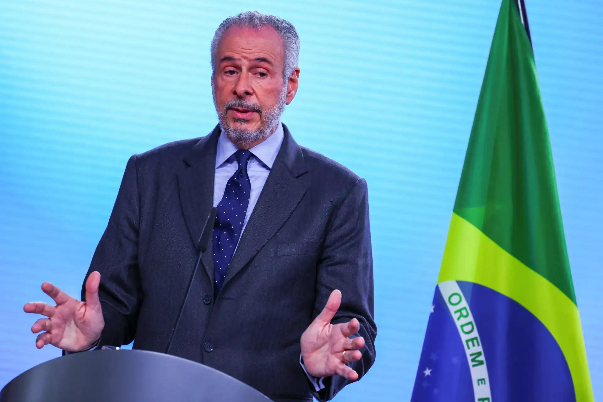 Homem de terno cinza com gravata, apresentando um discurso ao lado da bandeira do Brasil. Ele gesticula enquanto fala, demonstrando seriedade e envolvimento. A imagem transmite uma mensagem de liderança e responsabilidade. Ideal para temas relacionados à política e eventos oficiais no Brasil.