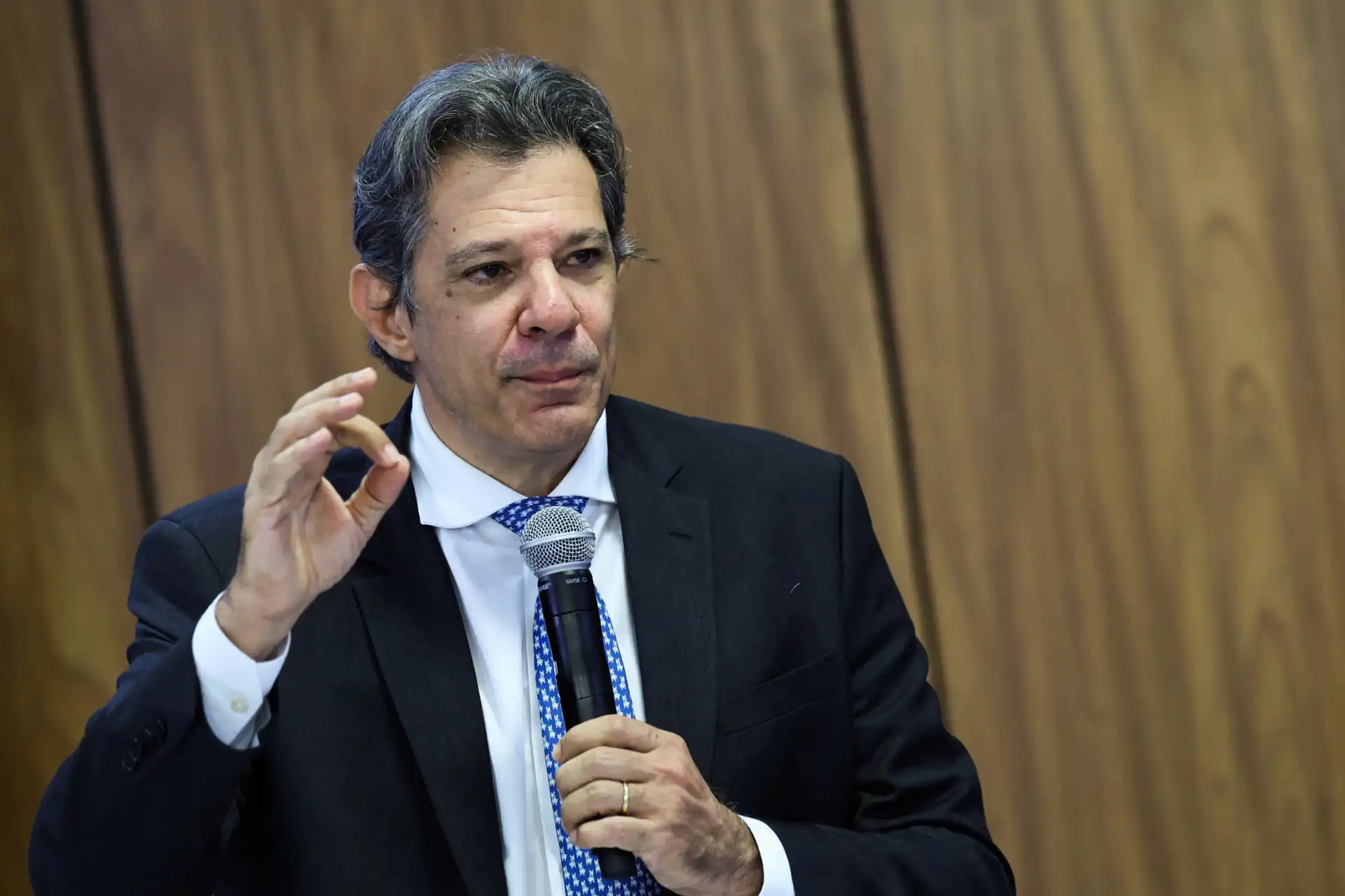 O político brasileiro Fernando Haddad faz um discurso, gesticulando com as mãos e segurando um microfone. Ele está usando um terno escuro com uma gravata azul estampada em um evento.