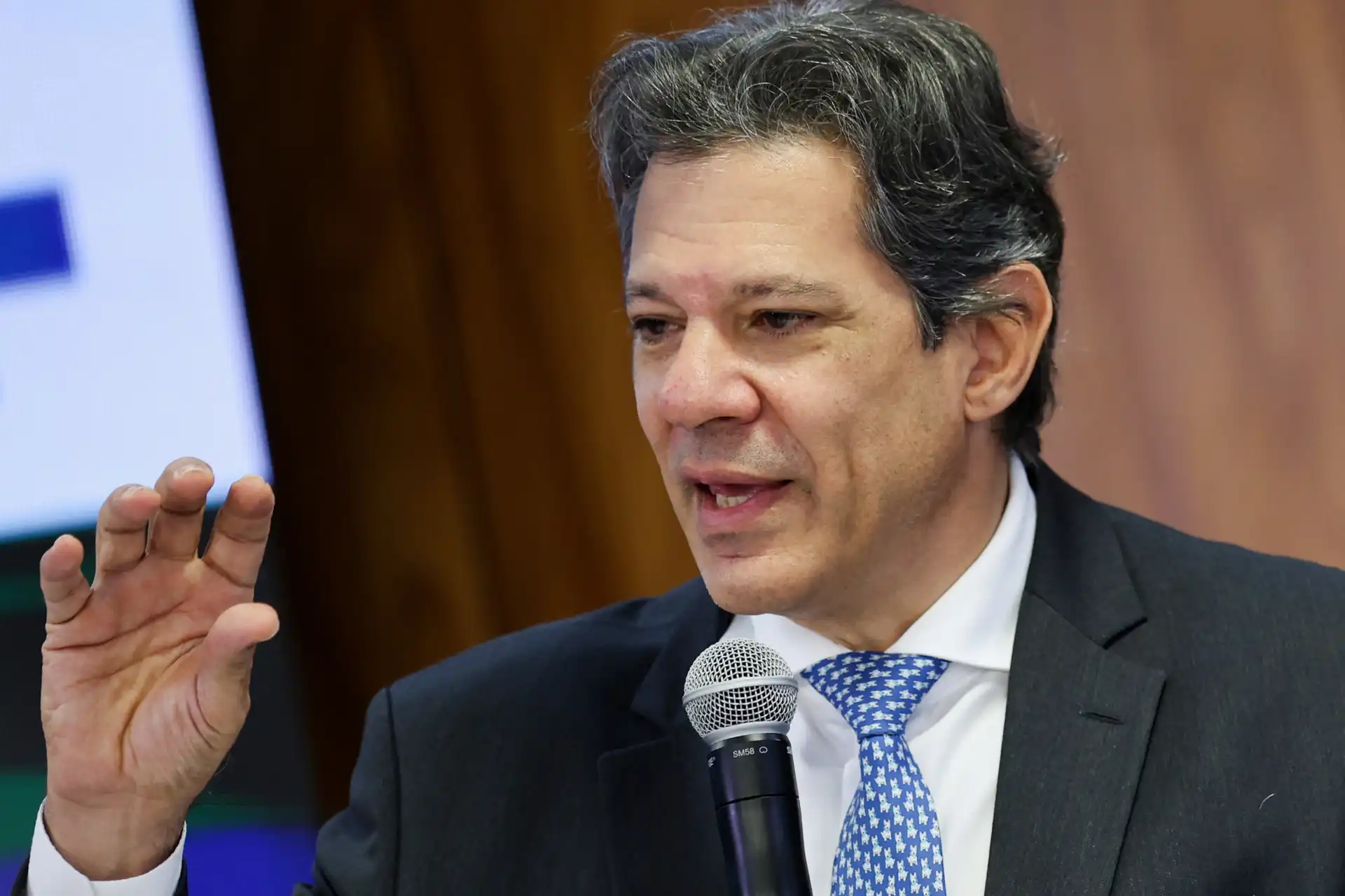 Haddad discursa em evento com microfone diante de uma tela, destacando suas propostas e visões políticas. Ele é conhecido por sua atuação como político e economista no Brasil.