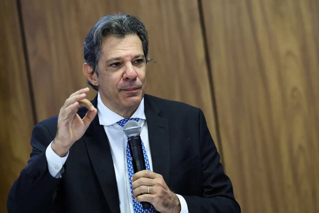 Homem de terno escuro, cabelo grisalho e expressão séria falando ao microfone em evento ou conferência, com fundo de painel de madeira