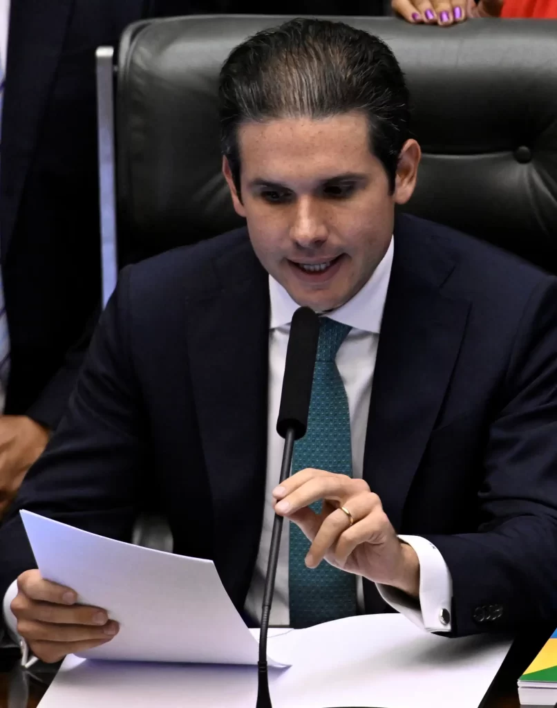 Câmara quer avançar com "pacote antifraude" após denúncias no INSS Um homem vestido com um terno elegante, sentado em uma mesa, segurando um documento e falando no microfone, em um ambiente formal.