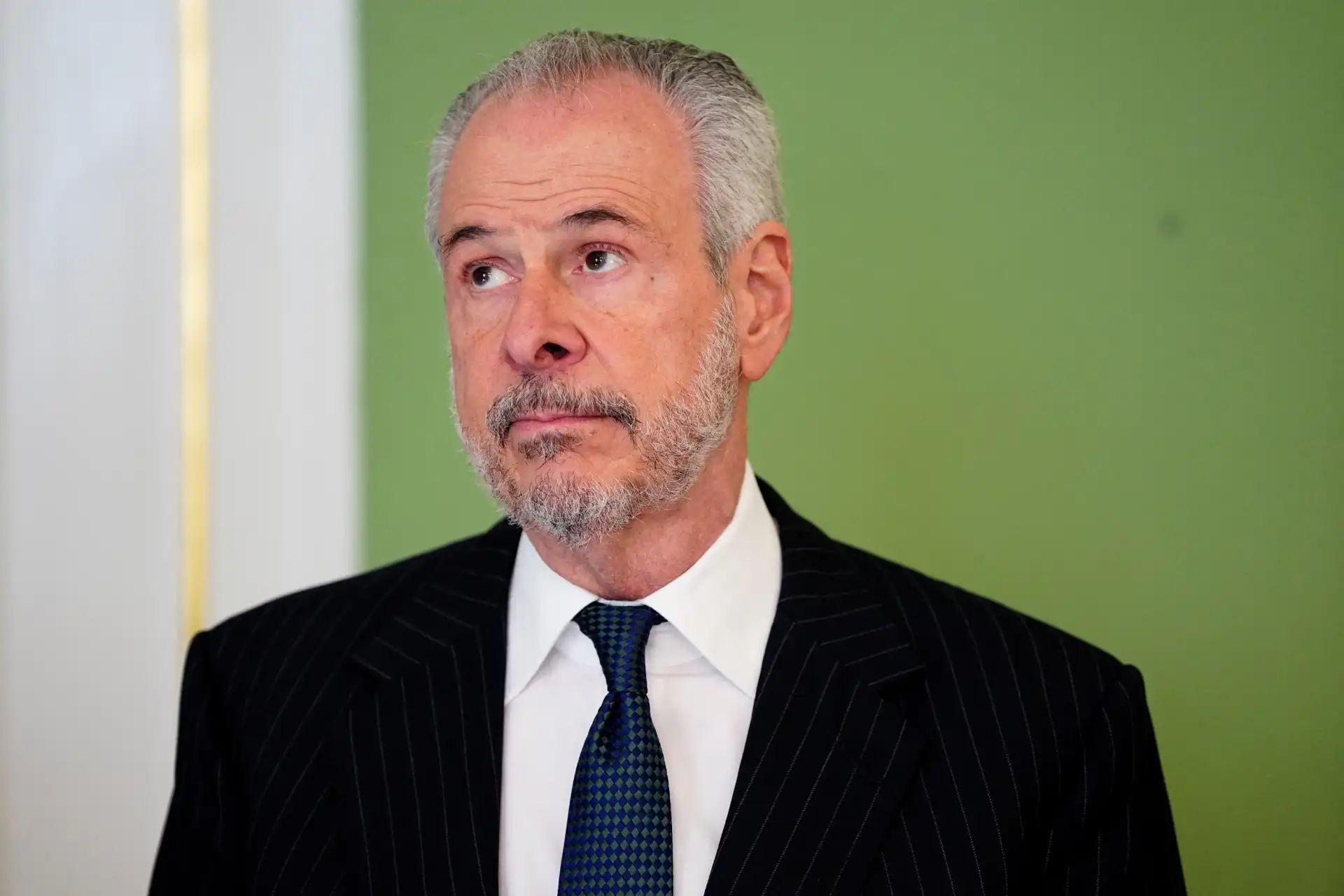 Homem de terno listrado em fundo verde, com expressão pensativa e cabelo grisalho, refletindo um momento de introspecção ou consideração.