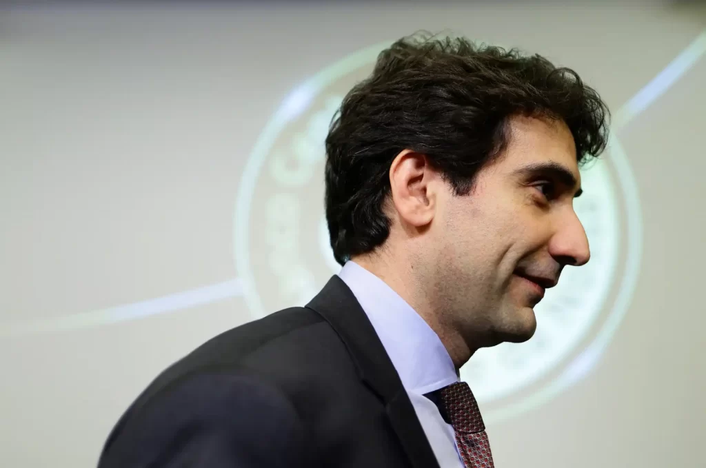 Descrição de um homem com cabelo escuro, usando um terno, observado de perfil em um ambiente formal. É uma imagem que evoca profissionalismo e foco.