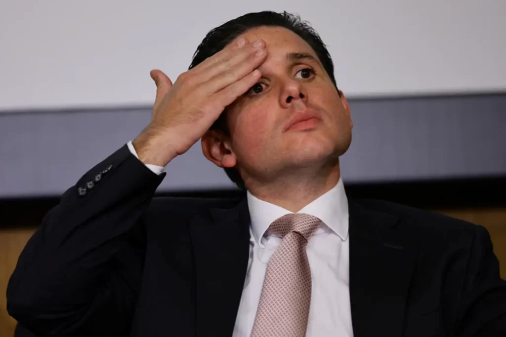Homem em traje formal demonstrando expressão de reflexão ou preocupação durante evento. Ele é um exemplo de liderança no ambiente de negócios, abordando temas relevantes.