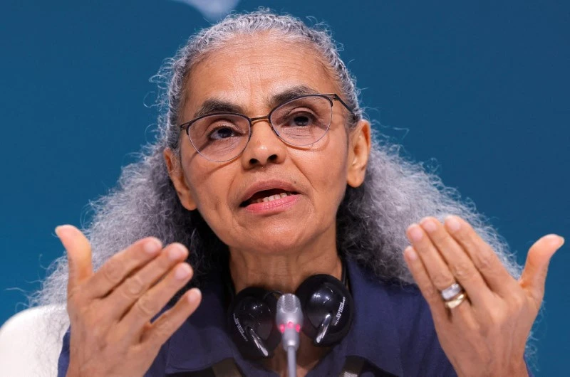 Imagem de uma mulher idosa com cabelos grisalhos, usando óculos, falando em um microfone durante uma palestra ou entrevista, com fundo azul.