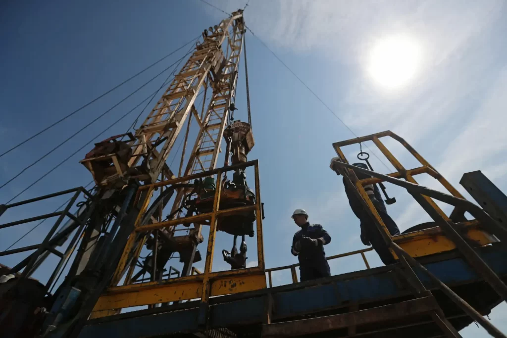 Operários trabalhando em plataforma elevada de uma torre de perfuração de petróleo sob céu claro, realizando manutenção em ambiente industrial.