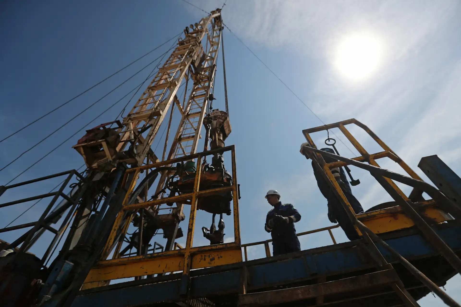 Operários trabalhando em plataforma elevada de uma torre de perfuração de petróleo sob céu claro, realizando manutenção em ambiente industrial.