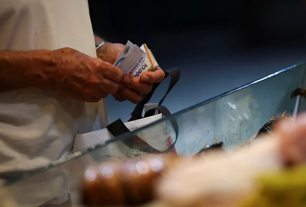 Pessoa segurando notas de dinheiro na fila do caixa de uma loja, com foco nas mãos e nas cédulas de real brasileiro, em ambiente de compras.