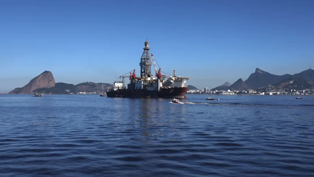 Imagem de uma plataforma de petróleo no mar, com detalhes do oceano e montanhas ao fundo, sob céu azul claro.