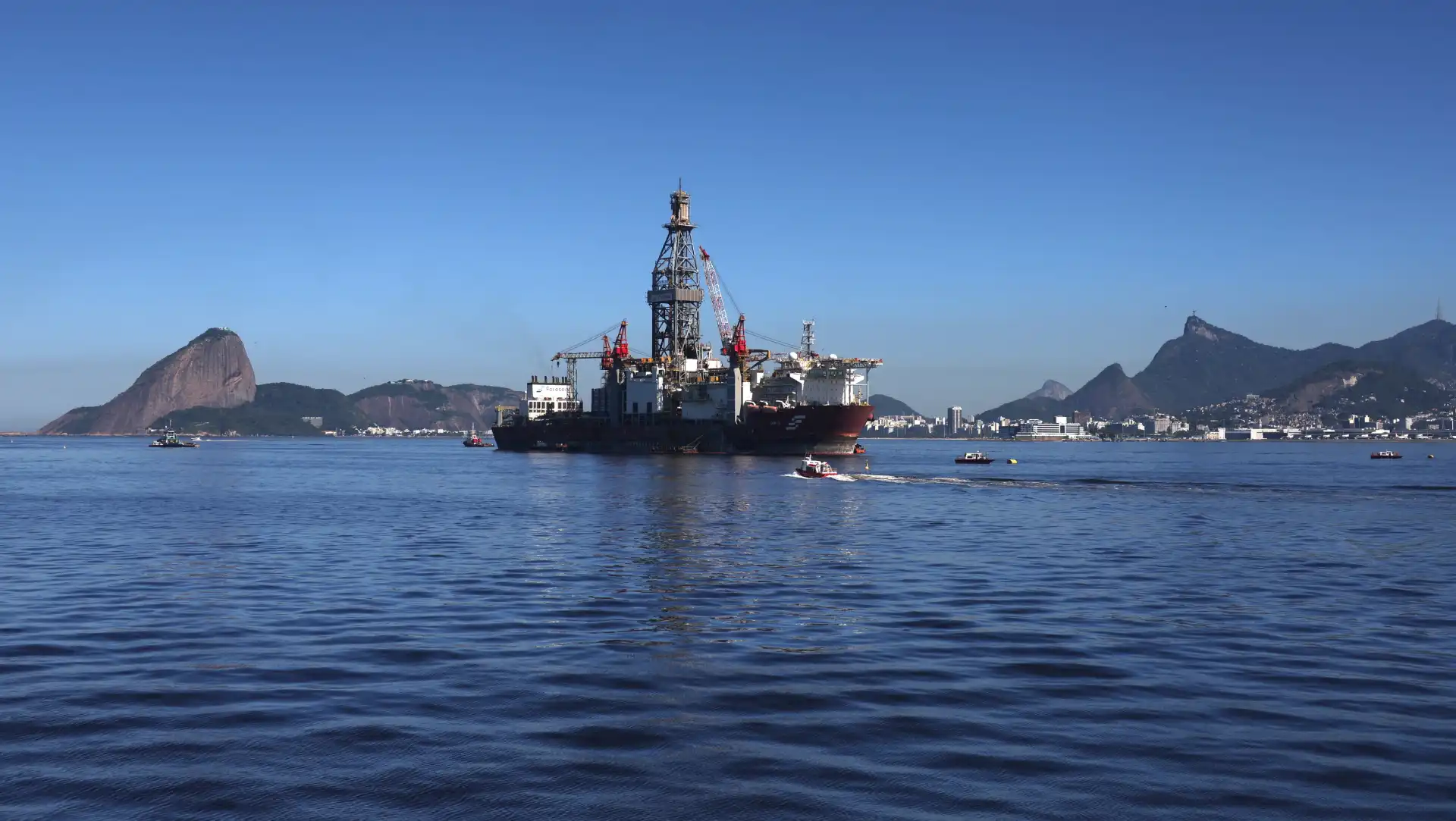 Imagem de uma plataforma de petróleo no mar, com detalhes do oceano e montanhas ao fundo, sob céu azul claro.