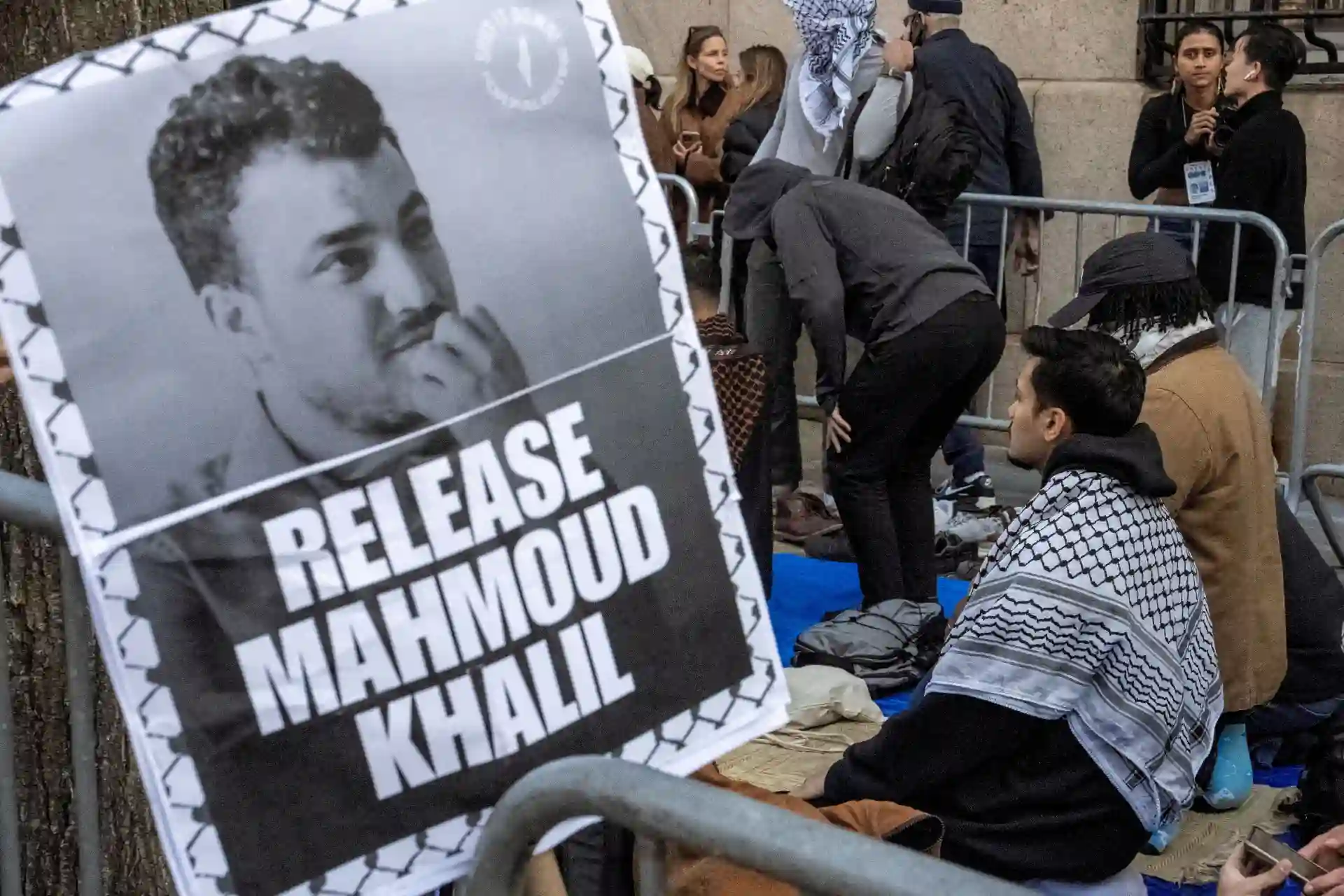 Imagem de um protesto pedindo a libertação de Mahmoud Khalil, com um cartaz em destaque. O fundo mostra pessoas reunidas, reforçando a manifestação.