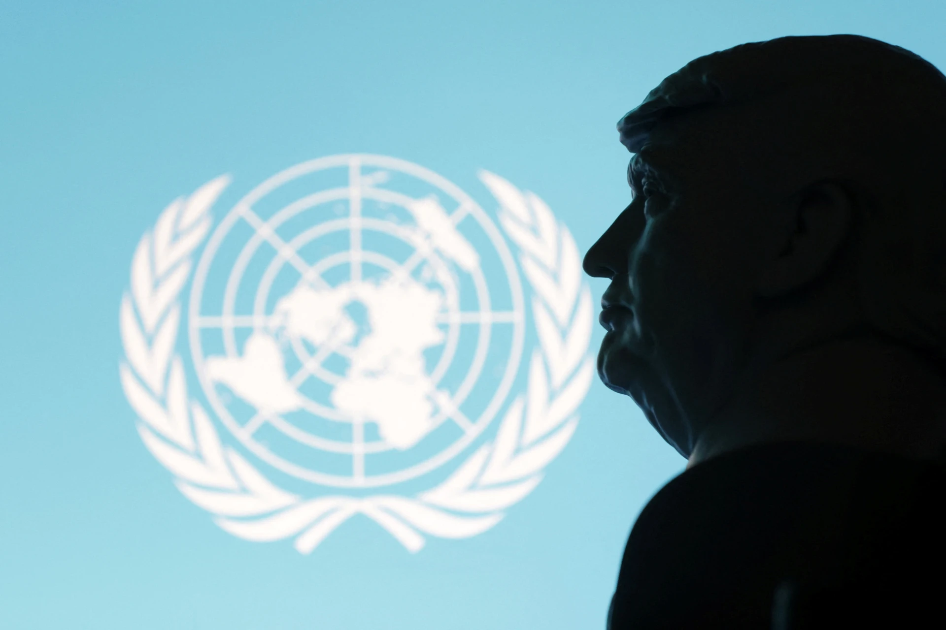Silhueta de uma figura em frente ao logotipo da ONU em um fundo azul claro, representando a importância da diplomacia e dos direitos humanos.