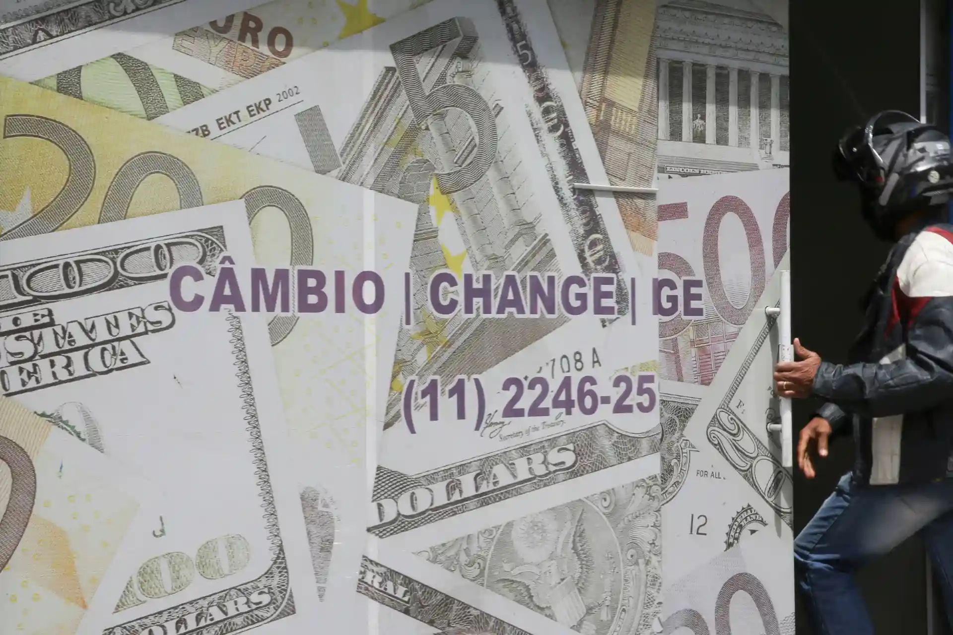 Pessoa caminhando ao lado de um mural com várias notas de dólares e euros, com destaque para a palavra Cambio em português, indicando troca de moedas.