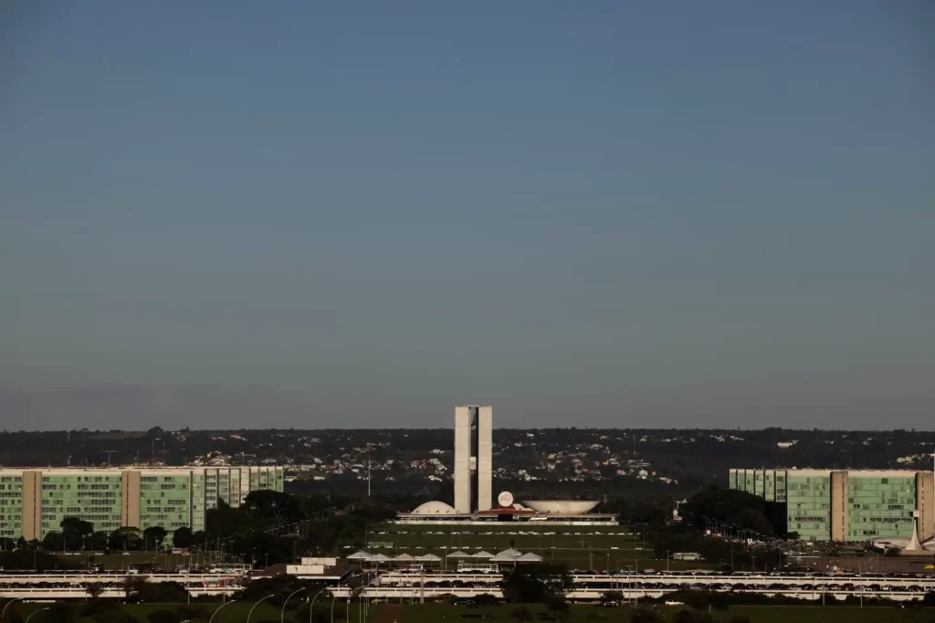 Vista panorâmica do Congresso Nacional em Brasília, com o icônico Palácio do Planalto e as torres de vidro ao fundo, sob um céu claro.