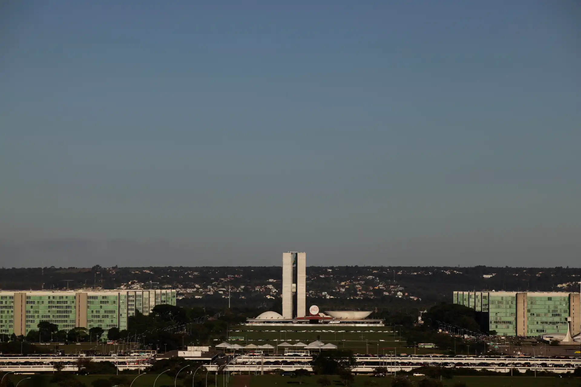 Vista panorâmica do Congresso Nacional em Brasília, com o icônico Palácio do Planalto e as torres de vidro ao fundo, sob um céu claro.