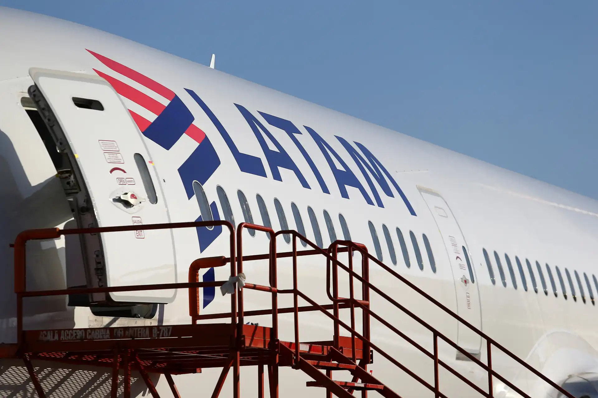 Imagem de aeronave da Latam Airlines em solo, com escada de embarque ao lado, destaque para o logotipo da companhia aérea e fuselagem branca sob céu claro.