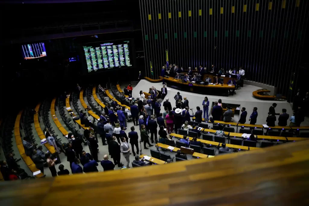 Sessão da assembleia legislativa da câmara com deputados e funcionários discutindo assuntos políticos e legislativos em plenário com destaque para o painel de votação digital ao fundo.
