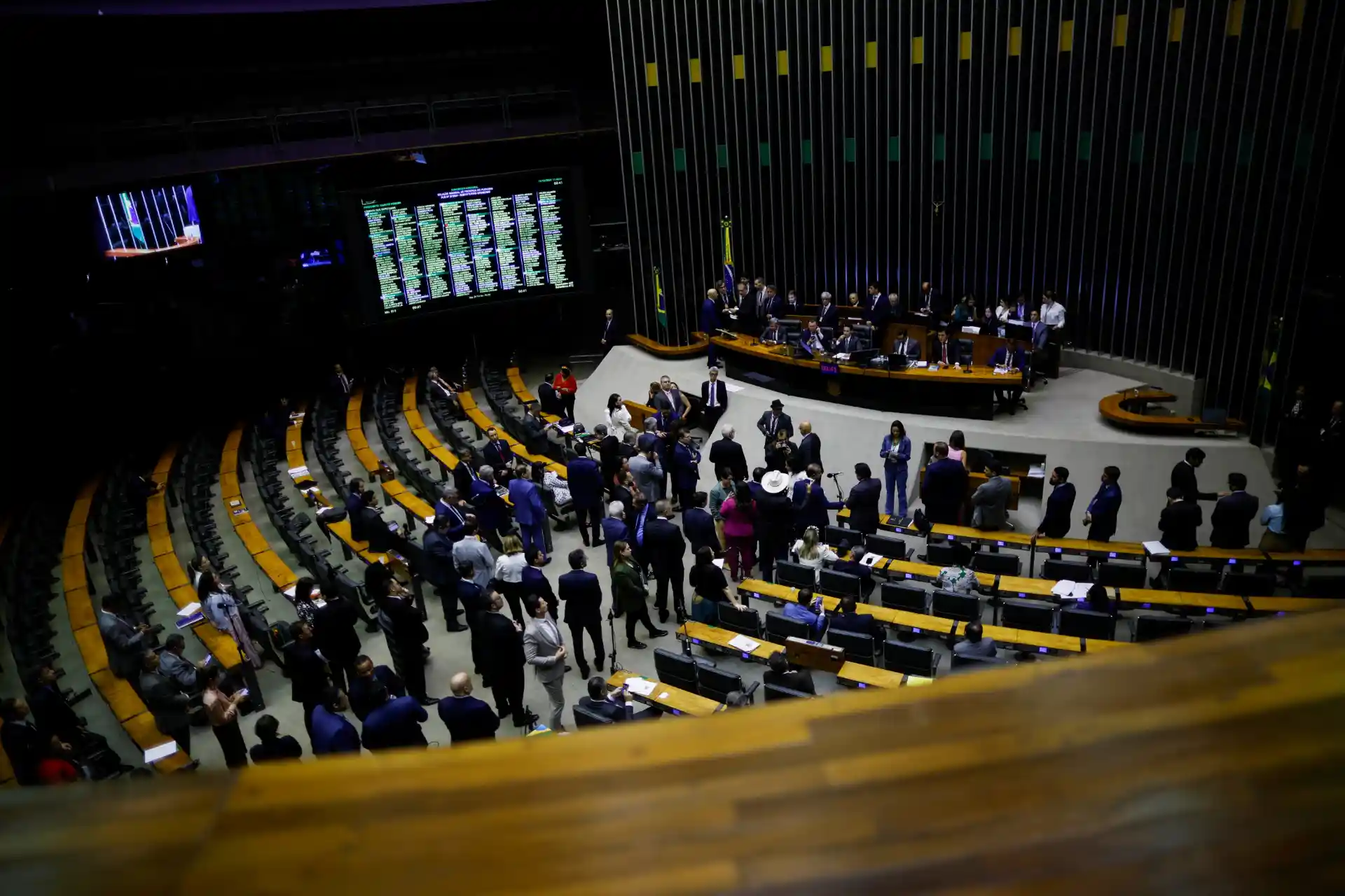Sessão da assembleia legislativa da câmara com deputados e funcionários discutindo assuntos políticos e legislativos em plenário com destaque para o painel de votação digital ao fundo.