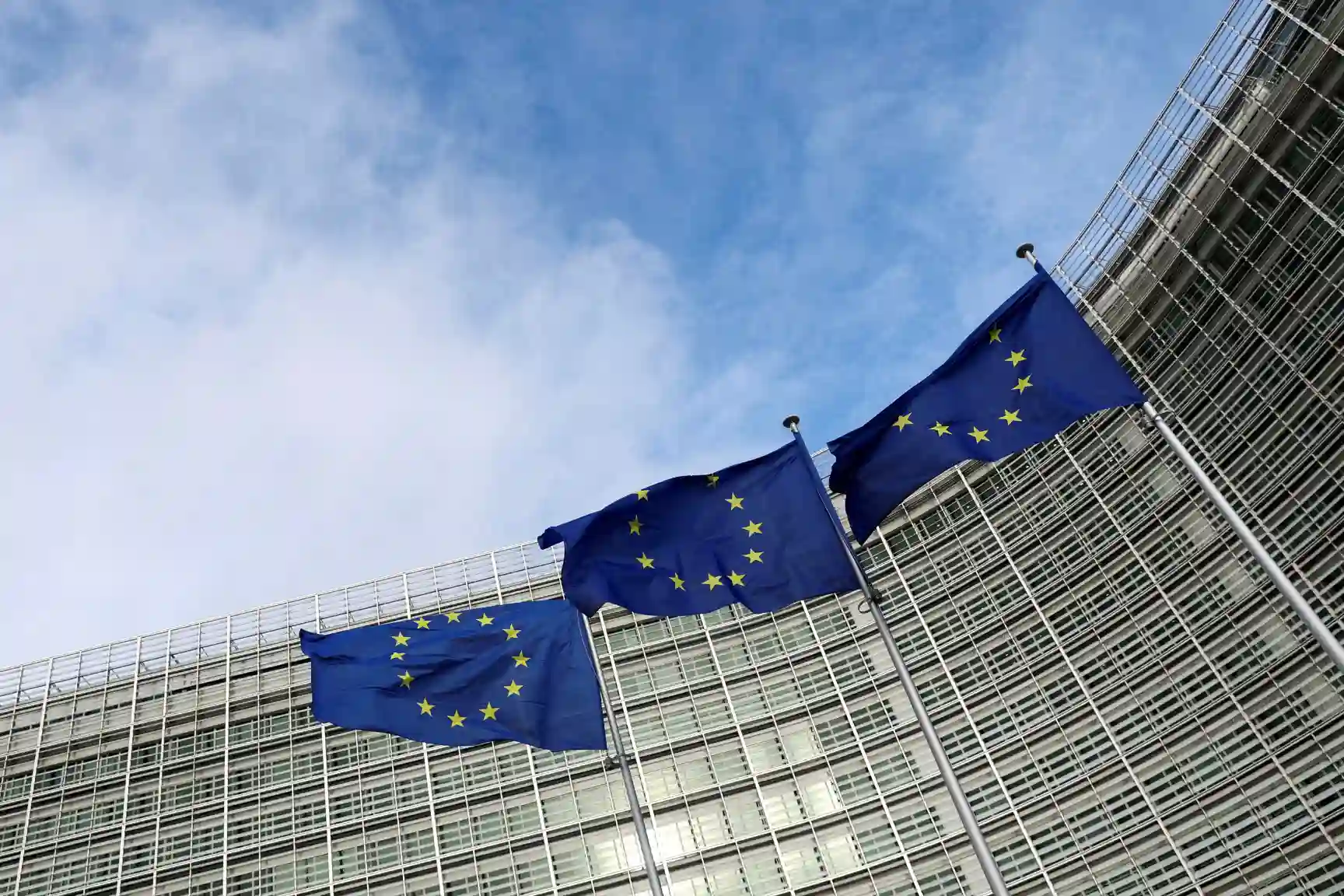 Três bandeiras da União Europeia agitadas ao vento na fachada de um edifício moderno com estrutura de vidro, sob céu azul com nuvens leves.