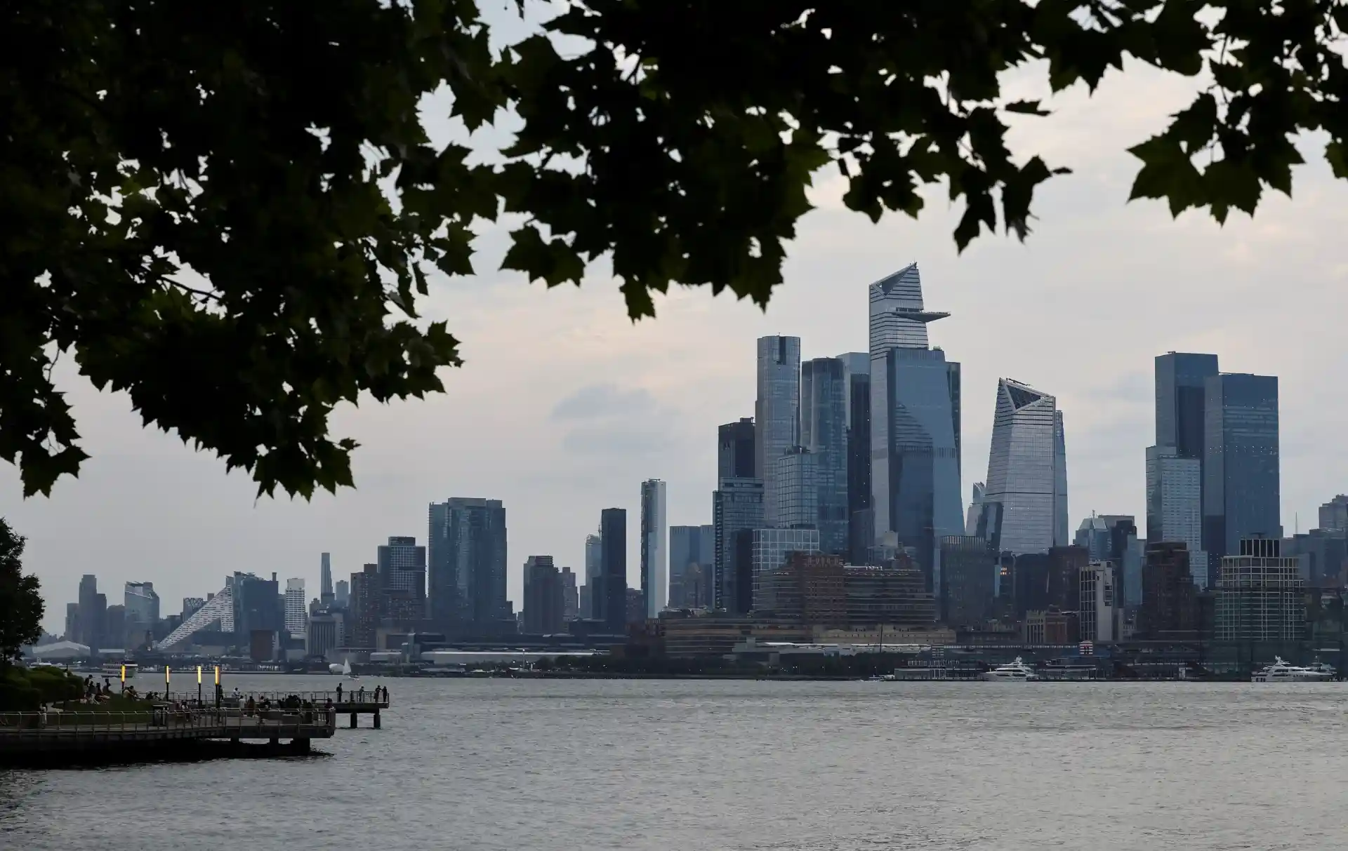 Vista da cidade de Nova York com arranha-céus modernos ao pôr do sol, vistos do rio, com árvores em primeiro plano para uma composição natural.
