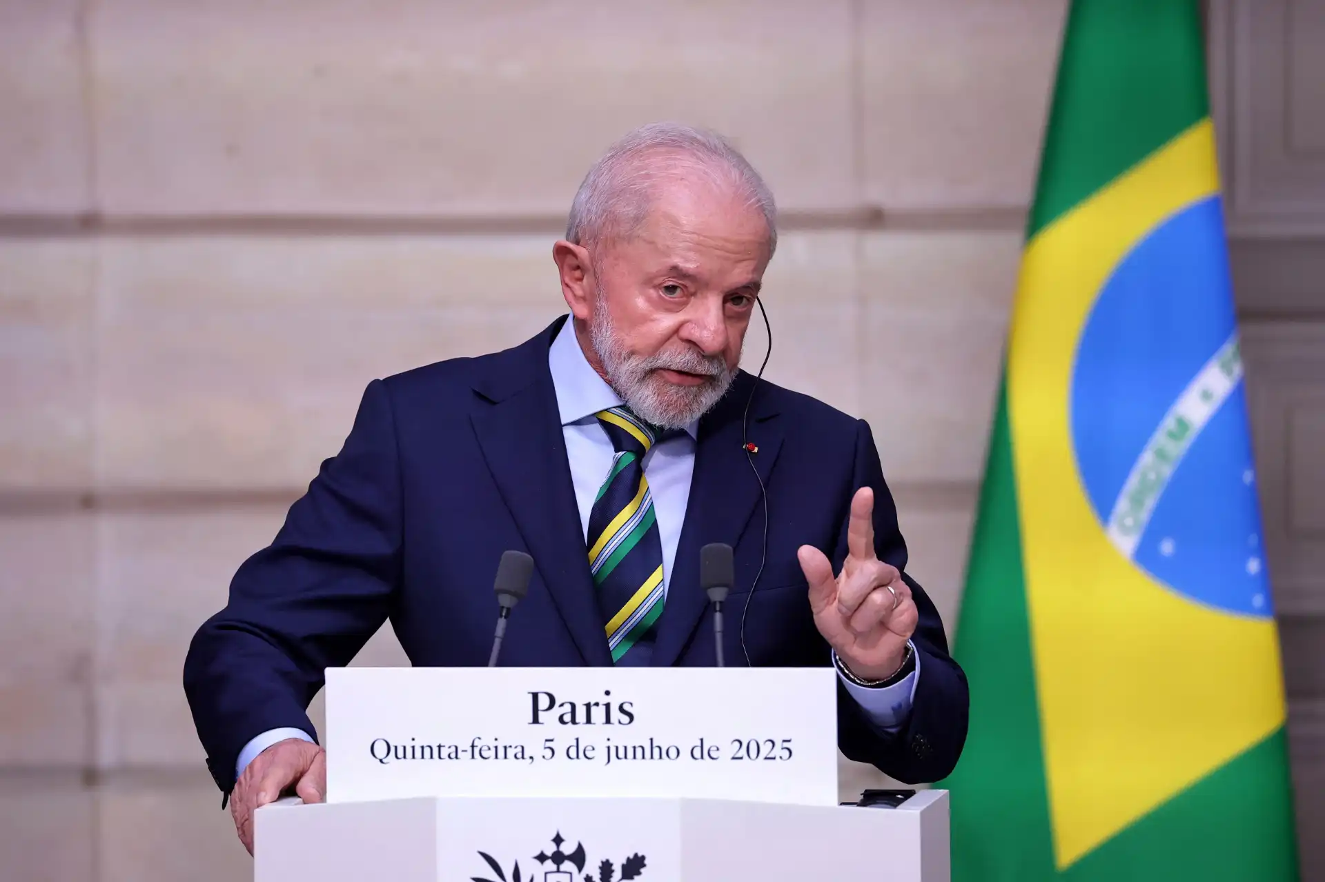 Imagem de um homem de terno azul escuro, barba branca e cabelos grisalhos, falando em um evento oficial ao lado da bandeira do Brasil, com um cartaz que mostra 'Paris, Quinta-feira, 5 de junho de 2025'.