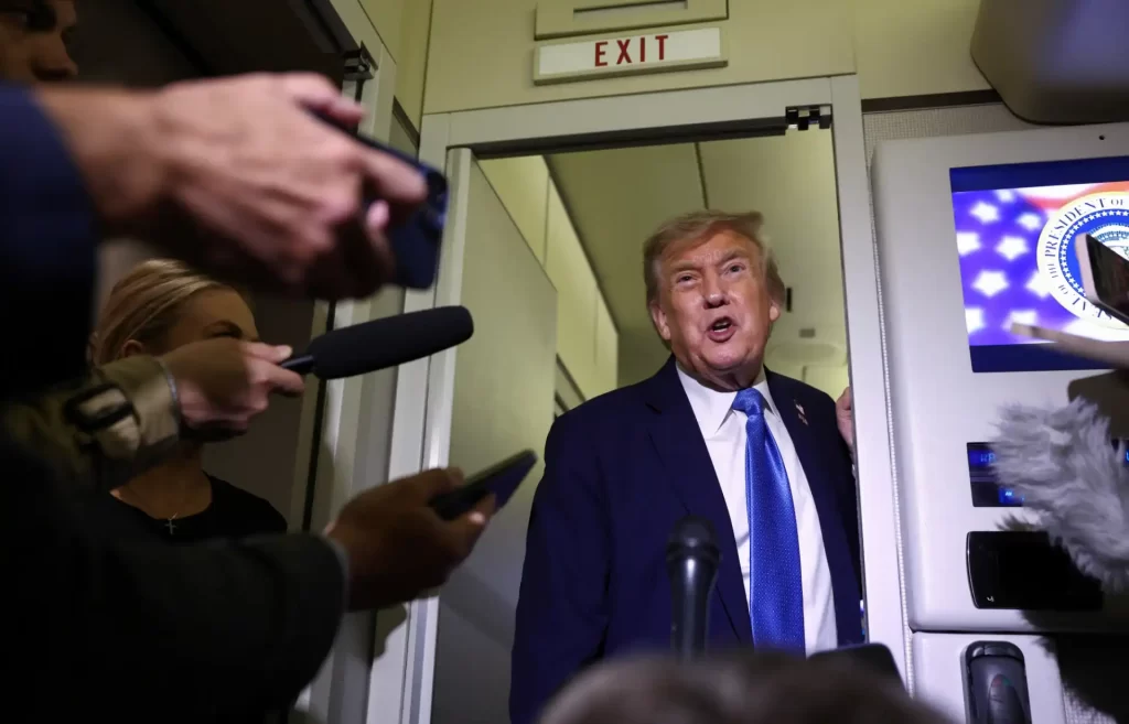 Imagem de Donald Trump, ex-presidente dos Estados Unidos, em uma entrevista ou declaração pública, rodeado por jornalistas com celulares e microfones, no interior de um prédio com sinal de saída. A cena mostra Trump falando e sendo registrado por diversos meios de comunicação.