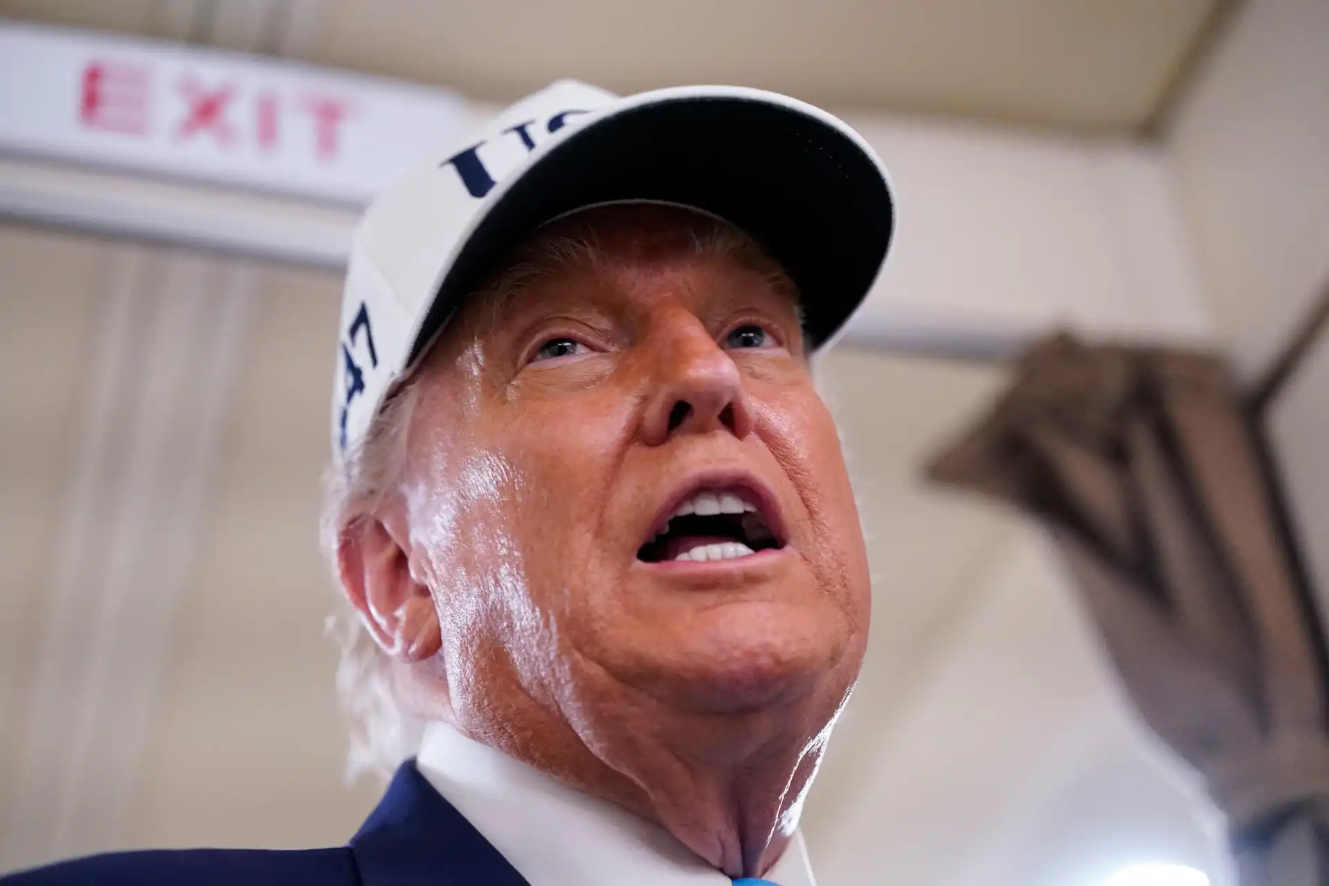 Imagem de Donald Trump durante um evento, com expressão facial intensa e expressão de fala, usando boné branco com inscrição.
