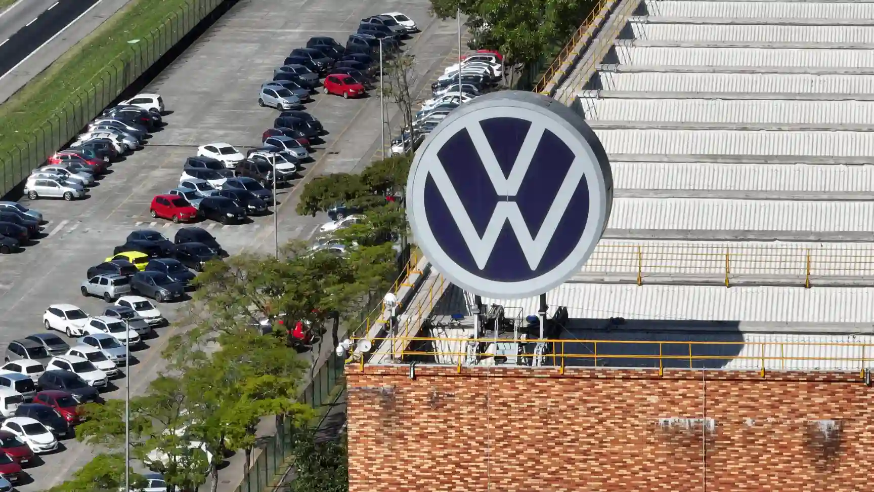 Estacionamento de veículos ao ar livre ao lado de uma loja Volkswagen, com logo da marca visível na imagem, em uma área urbana com árvores e edifícios.