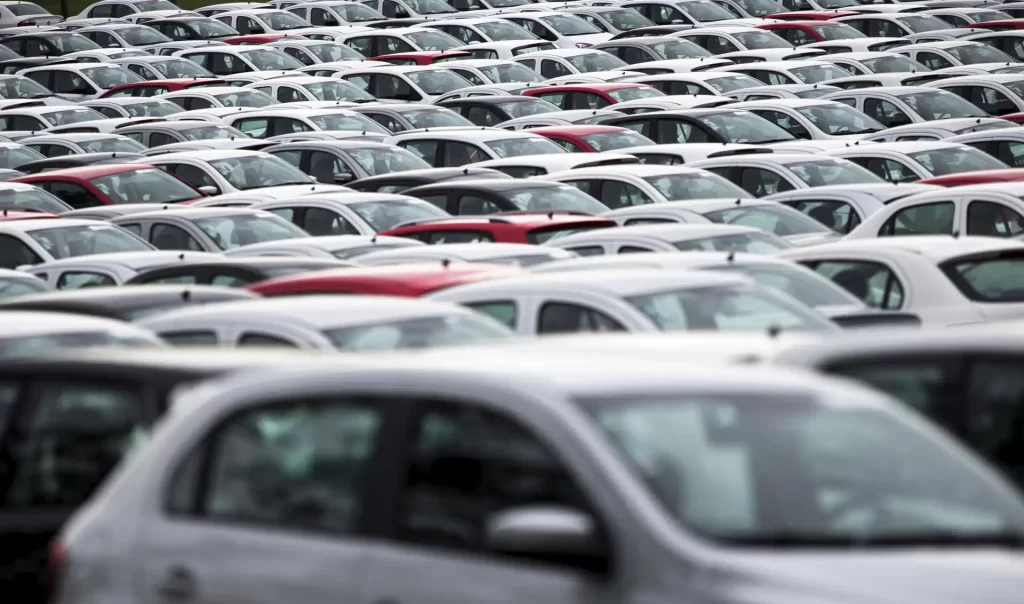 Imagem de um grande estacionamento cheio de carros brancos e alguns vermelhos, mostrando a vastidão de veículos estacionados em um espaço amplo.