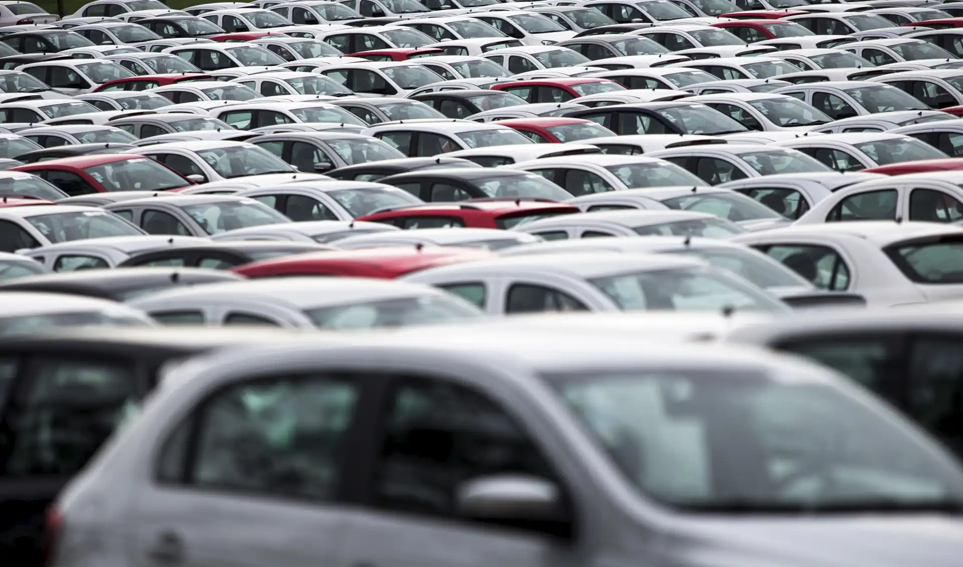 Imagem de um grande estacionamento cheio de carros brancos e alguns vermelhos, mostrando a vastidão de veículos estacionados em um espaço amplo.