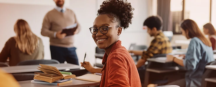 Estudante afrodescendente com óculos sorrindo em sala de aula, interagindo com colegas e professor, ambiente de aprendizagem estimulante e inclusivo.