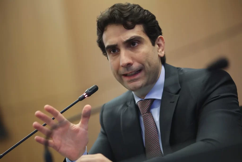 Homem em traje formal falando em uma reunião ou conferência, gesticulando com a mão à frente de uma mesa.