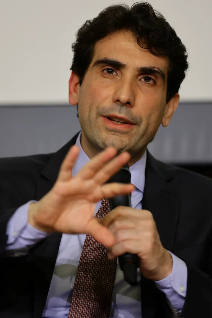 Homem de terno escuro e camisa clara, falando ao microfone durante uma coletiva ou apresentação, com expressão séria e gesticulando com a mão.