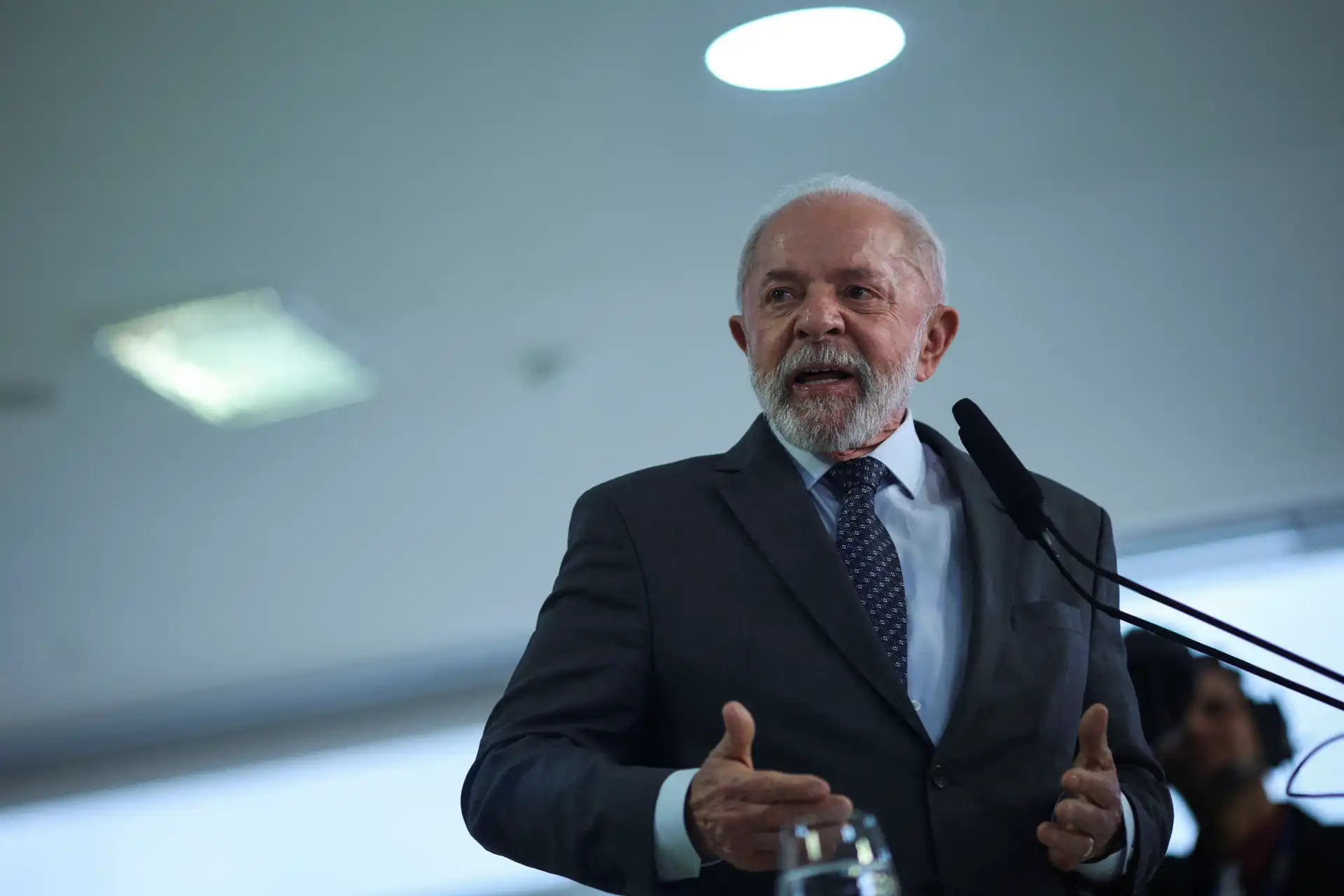 Homem idoso com barba branca e cabelo grisalho, vestido com terno escuro e gravata, fala em podium durante uma palestra ou evento, focado na palavra-chave.