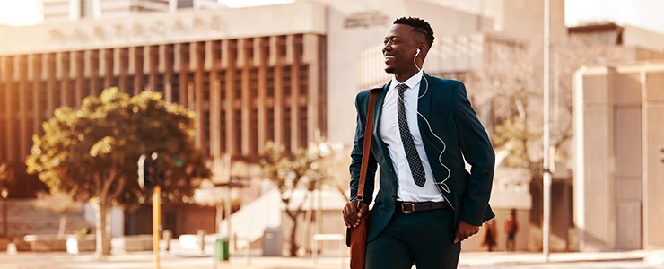 Homem de negócios sorridente caminhando na cidade com fones de ouvido e roupa formal ao sol, representando sucesso e empreendedorismo e liberdade de expressão