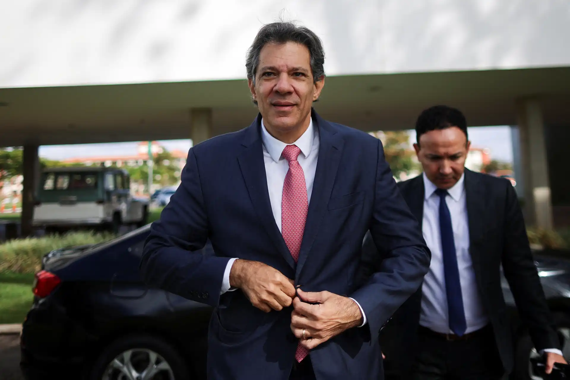 Homem político em um evento ao ar livre, vestindo terno azul com camisa branca e gravata rosa, com outro homem ao fundo, ao lado de um carro preto.