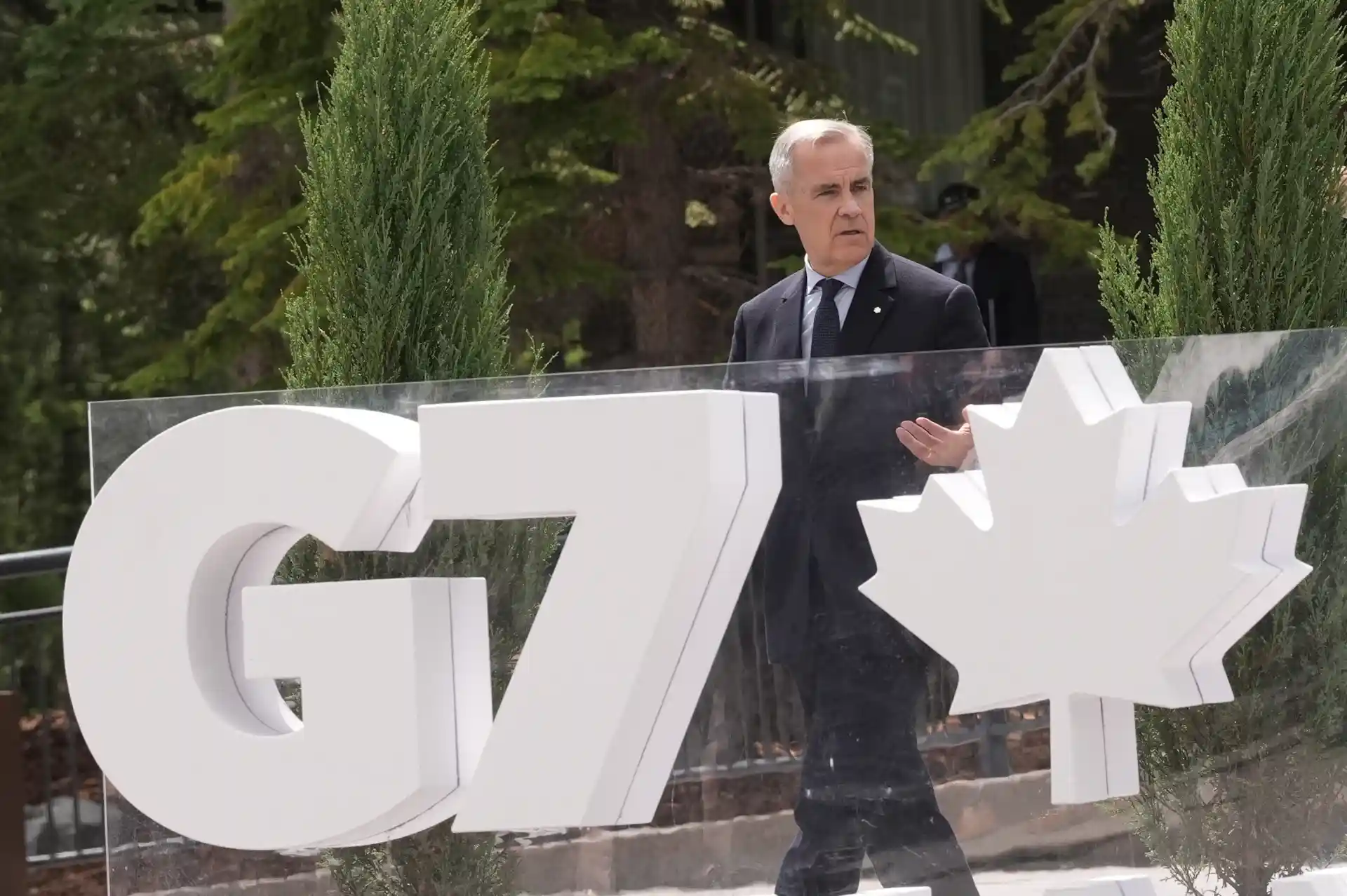 Homem de terno preto e camisa branca ao lado de símbolo do G7 e folha de maple, em ambiente externo com árvores verdes ao fundo.