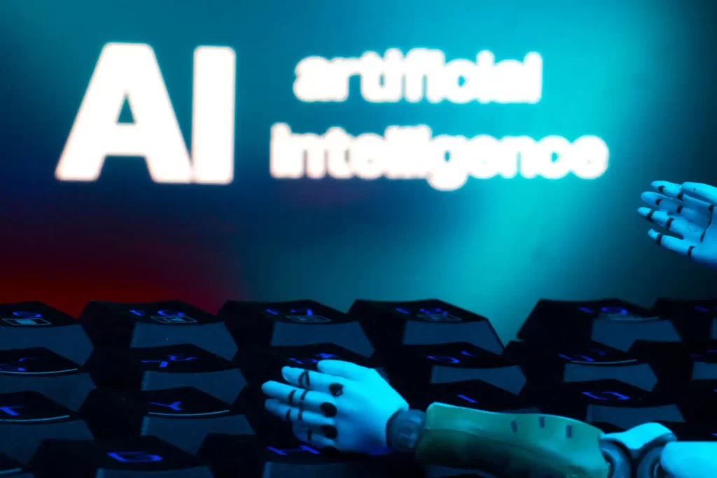 Imagem de um robô com mãos robóticas ao lado de computadores conectados, com destaque para o logo de inteligência artificial na tela ao fundo, representando tecnologia de IA e automação.