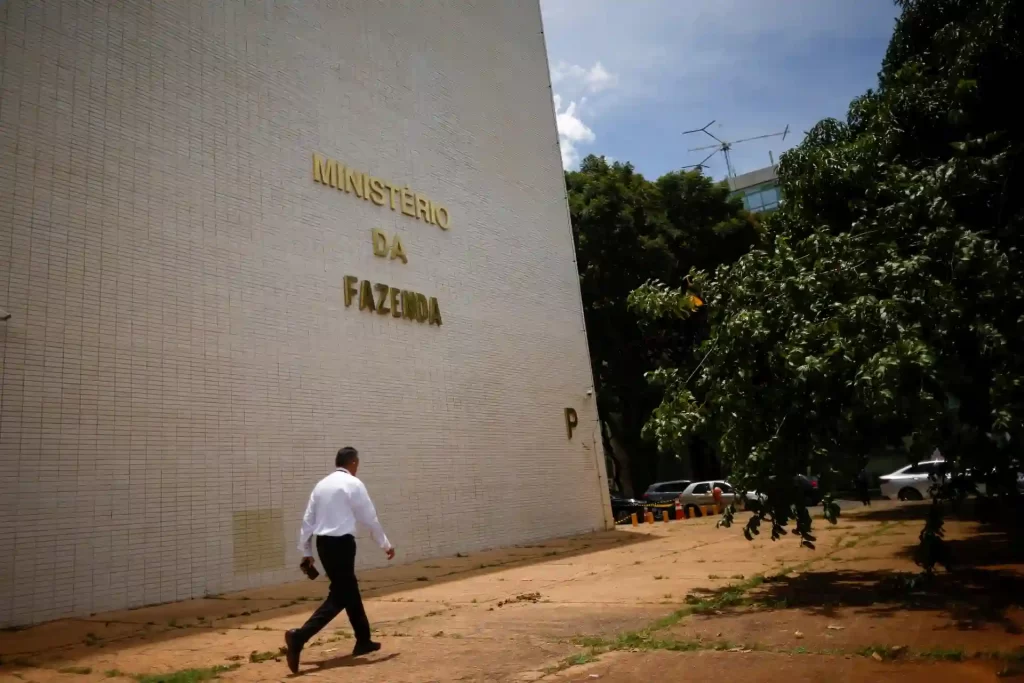 Imagem do edifício do Ministério da Fazenda com uma pessoa caminhando em frente, árvores e veículos ao fundo, sob céu parcialmente nublado, relacionado ao Ministério da Fazenda.